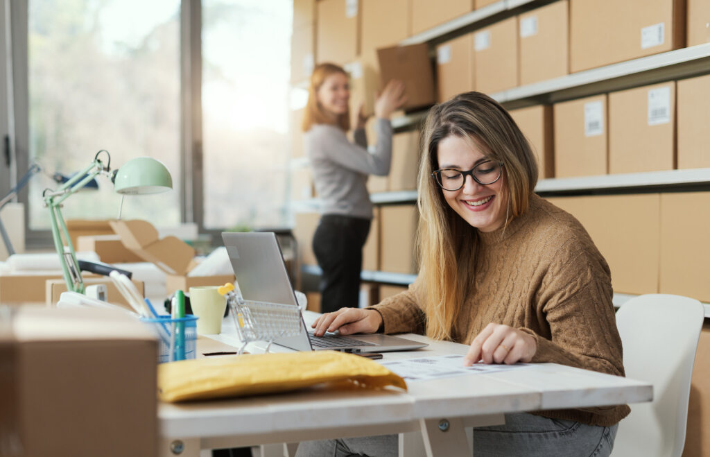 Deux femmes au travail, l'une floue à l'arrière-plan, rangeant une boîte en carton, et l'autre au centre de la photo, assise devant un ordinateur, souriante, regardant un document posé sur la table.