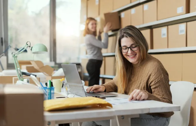 Deux femmes au travail, l'une floue à l'arrière-plan, rangeant une boîte en carton, et l'autre au centre de la photo, assise devant un ordinateur, souriante, regardant un document posé sur la table.
