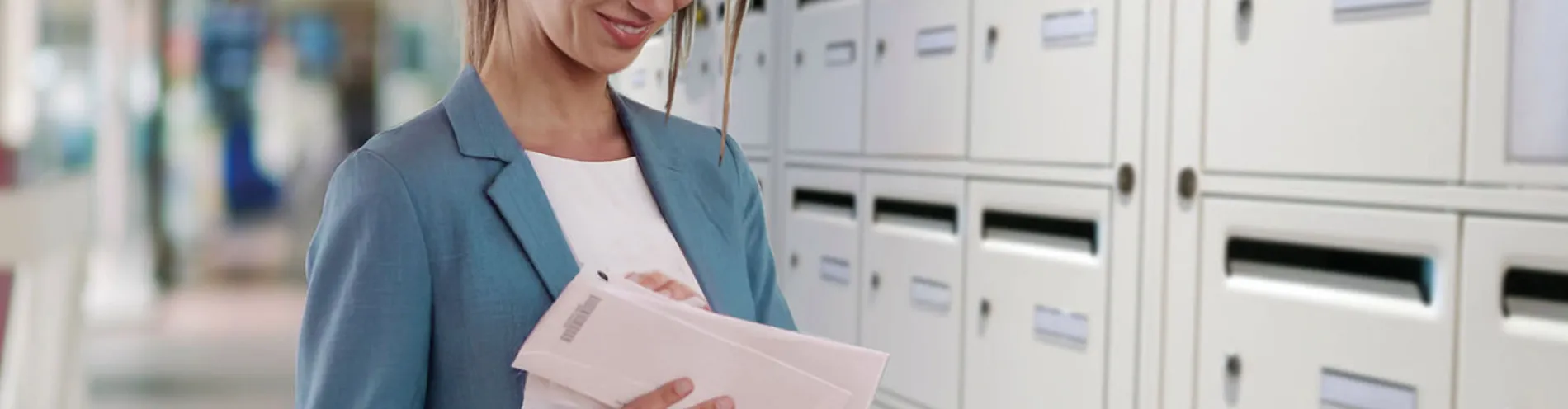 Une jeune femme brune consulte son courrier distribué par La Poste Solutions Business devant un mur de boîte à lettres 